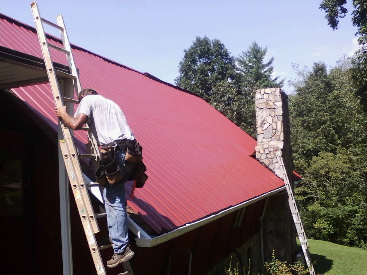 Roofer on a ladder beside a red metal roof and stone chimney — Soffit & Fascia Repair by Roofing Brothers in White Mills